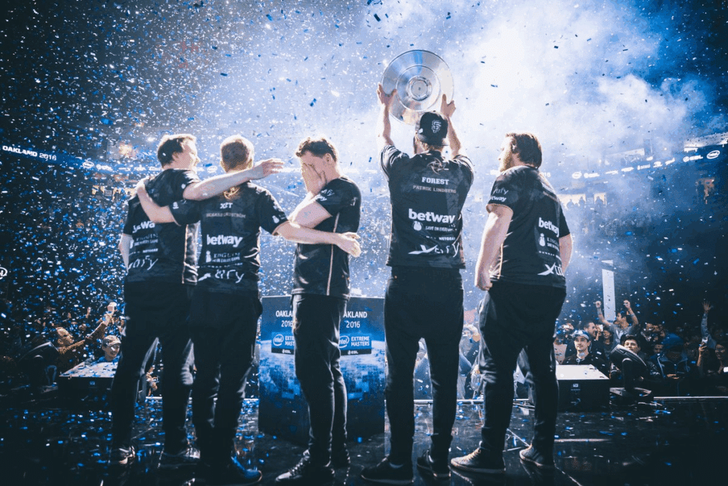 Esports team celebrating with a trophy on stage amidst a shower of confetti and cheering crowd