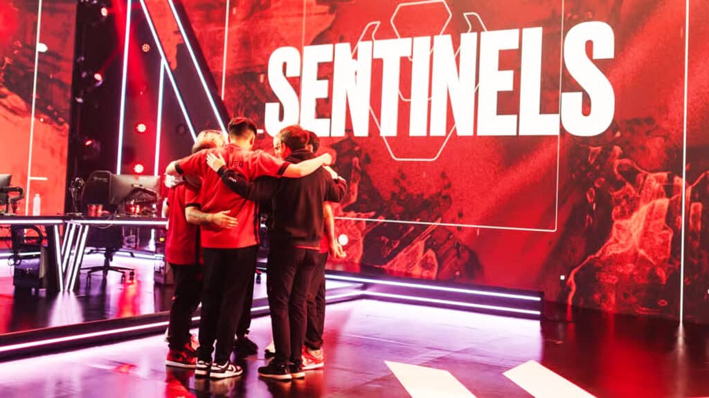 Esports team in red jerseys huddling on stage in front of a large Sentinels banner.