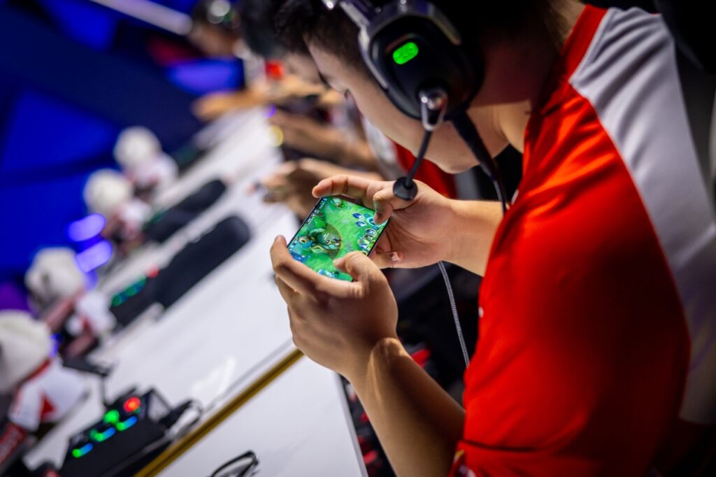 Gamer intensely focused on a mobile game during an esports event, wearing headphones, surrounded by gaming equipment